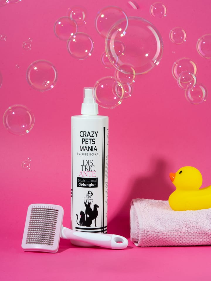 Still life su fondo fucsia attraversato da bolle di sapone: lo spray districante Crazy Pets Mania accanto a una spazzola cardatrice, un asciugamano rosa arrotolato e una paperella gialla