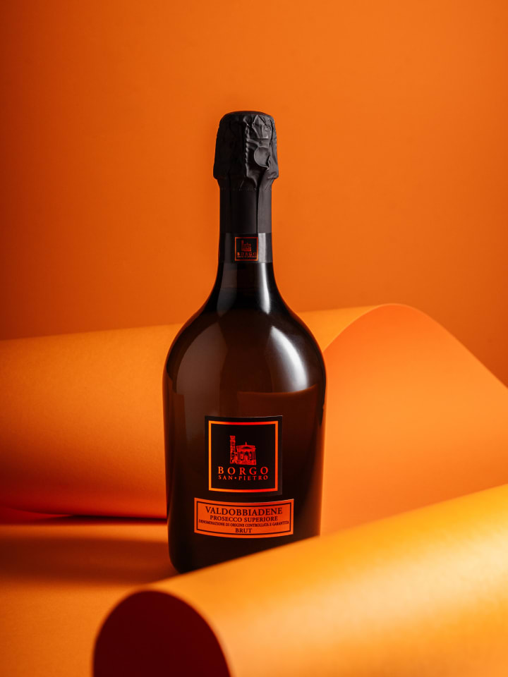 Bottiglia Borgo San Pietro Valdobbiadene Prosecco Superiore Brut su fondo arancione con fogli di carta arrotolati