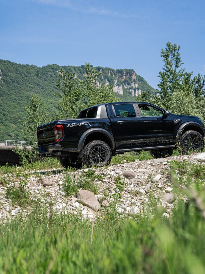 Ford Ranger Raptor nero di tre quarti posteriore su un greto di sassi, sotto le pareti di roccia della valle