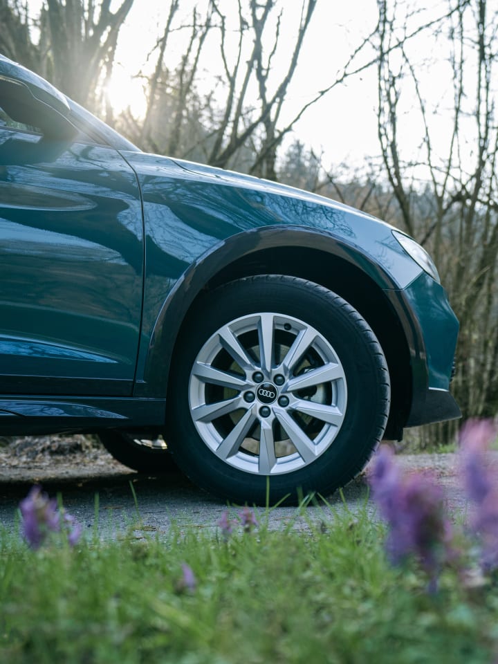 Ruota in lega e passaruota di un’Audi verde petrolio in controluce, con i fiori viola del prato in primo piano