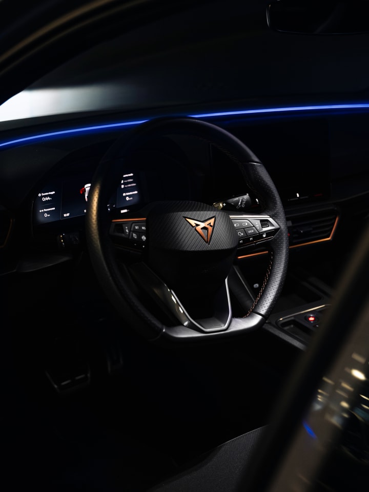 Volante di una Cupra con il logo in rame e la luce ambiente blu sul cruscotto, nell’abitacolo al buio