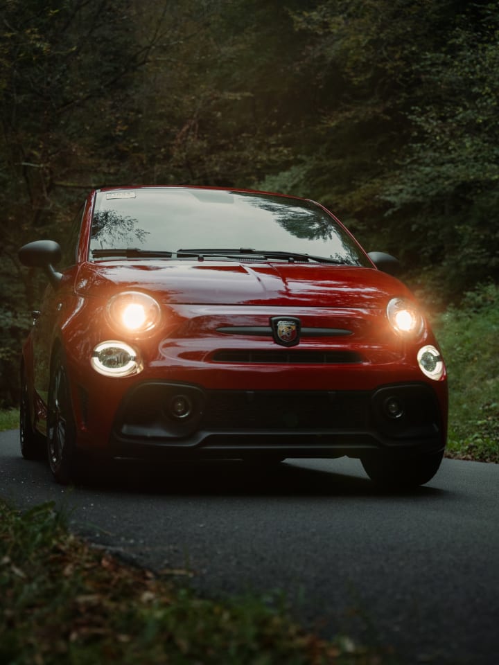 Abarth rossa con i fari accesi su una strada nel bosco, al calare della luce