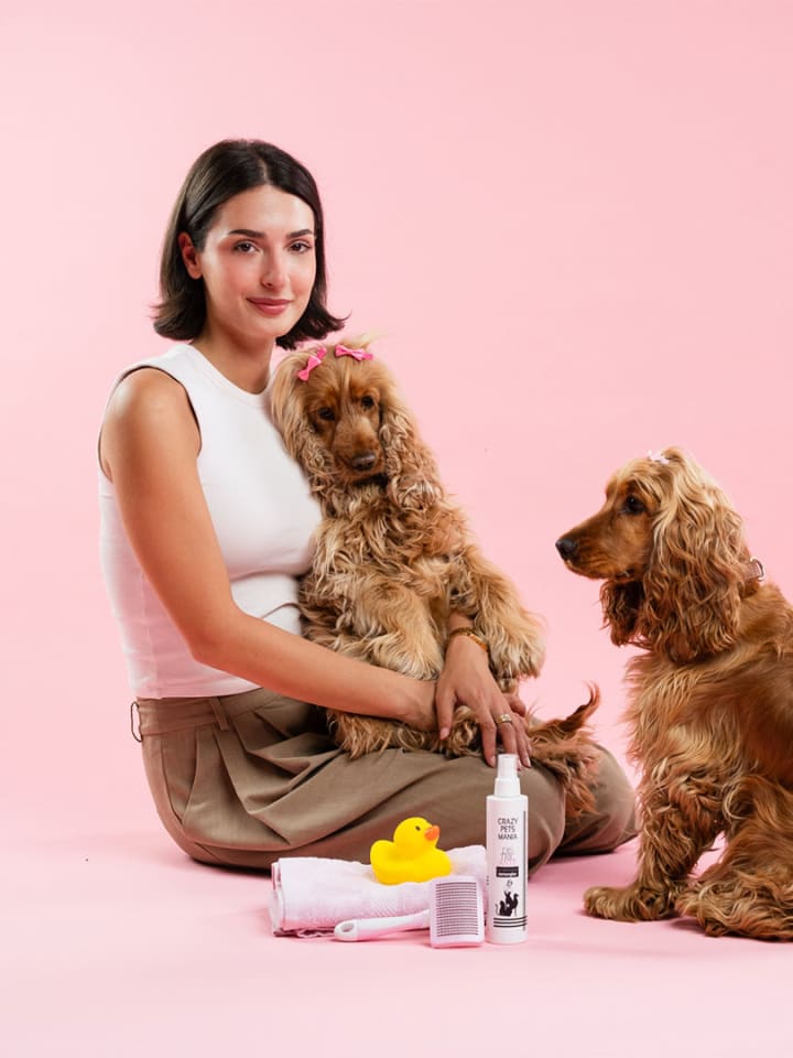 Ragazza seduta a terra su fondo rosa tra un cocker spaniel fulvo e un barboncino chiaro, con i flaconi Crazy Pets Mania e una paperella gialla accanto