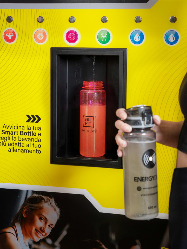 L’erogatore giallo All Fit per le Smart Bottle: una borraccia si riempie nella nicchia mentre una ragazza avvicina la propria, accanto alla scritta «Avvicina la tua Smart Bottle e scegli la bevanda più adatta al tuo allenamento»