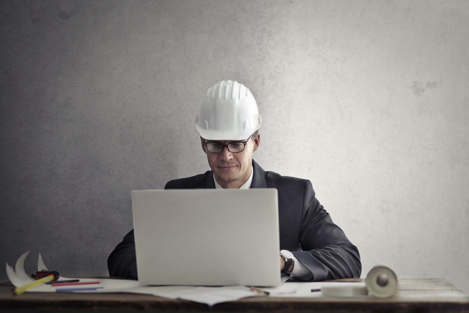 Serious professional male engineer in protective helmet and formal suit sitting at table and using laptop while working on project at workplace