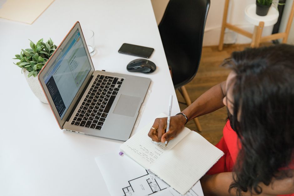 Businesswoman writing in notebook at office desk with laptop and plant.