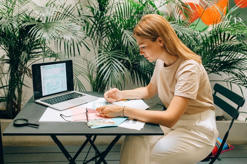 A woman deeply concentrated on work at her laptop amidst indoor greenery and documents.