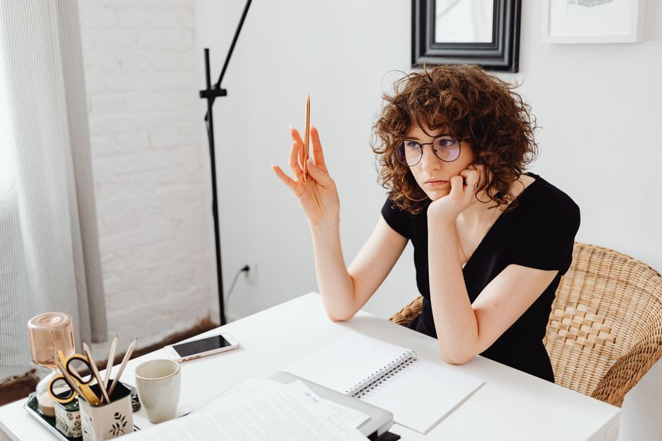 Young woman with curly hair in glasses thinking at her desk with a notebook, indoors.