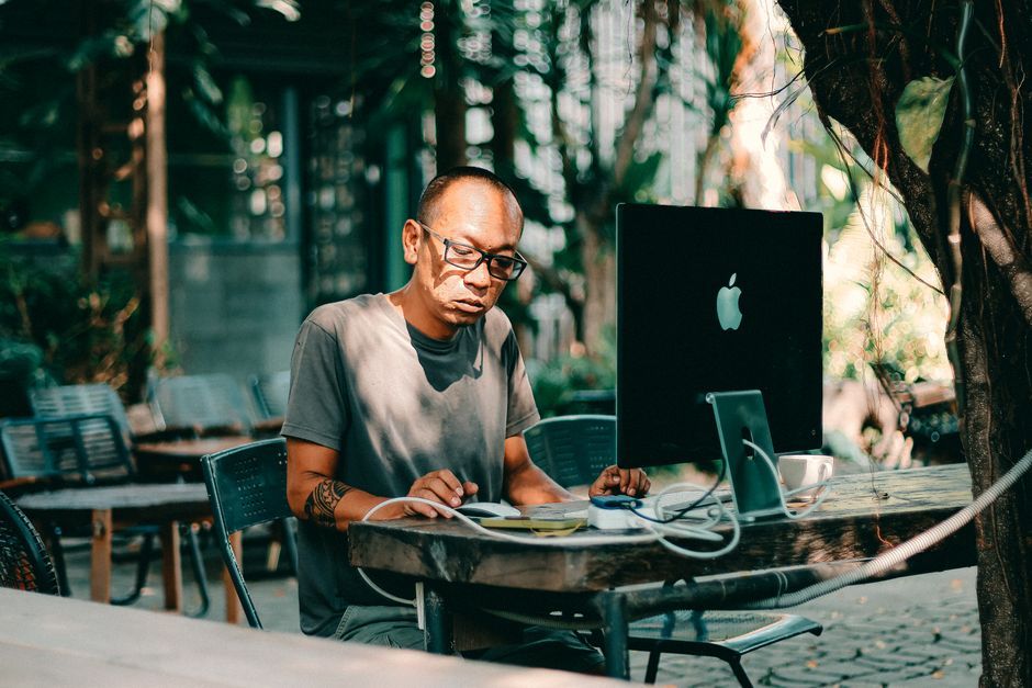 Adult man with glasses working on a computer outdoors in a sunny cafe setting.
