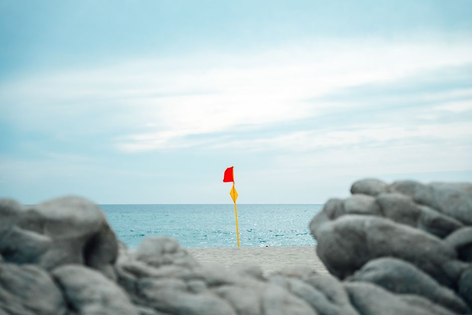 A red warning flag on a sandy beach in Oaxaca, Mexico, with ocean and rocky foreground.