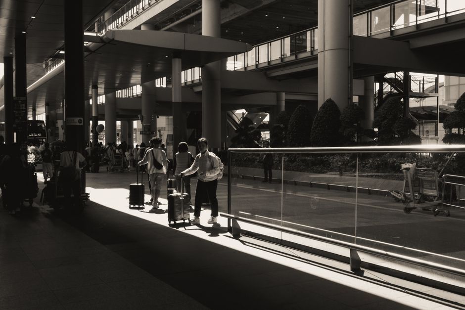 Silhouettes of travelers with luggage at a sunlit modern airport terminal in black and white.