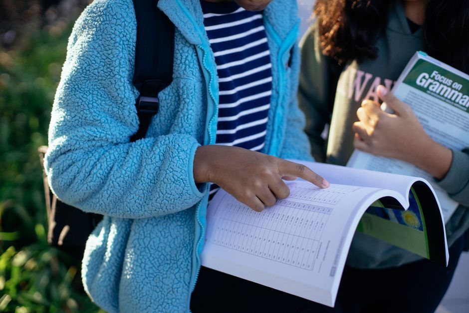 Crop anonymous ethnic schoolgirls pointing at exercise book page while studying together in sunlight