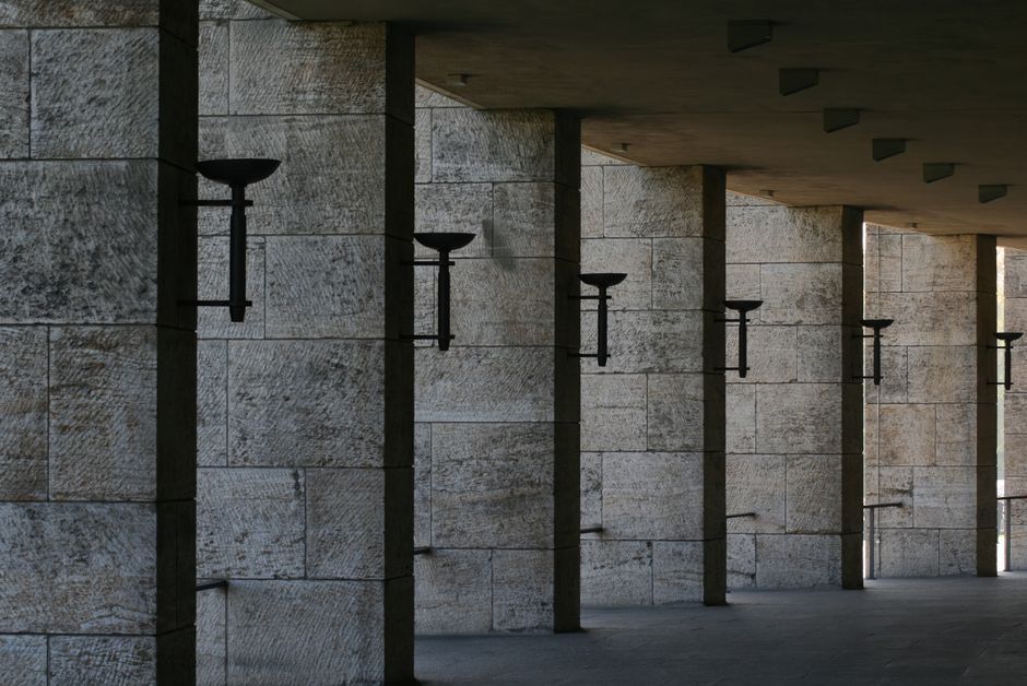A perspective view of a stone colonnade with torch holders casting shadows in an urban environment.