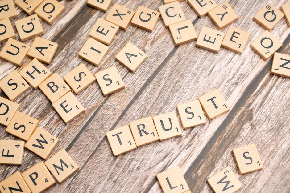 Scrabble letter tiles form the word 'Trust' on a wooden table, surrounded by scattered tiles.