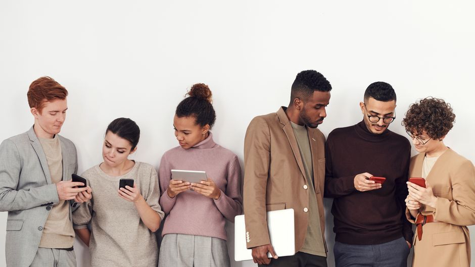Five adults in casual wear using phones and tablets indoors, emphasizing technology and fashion.