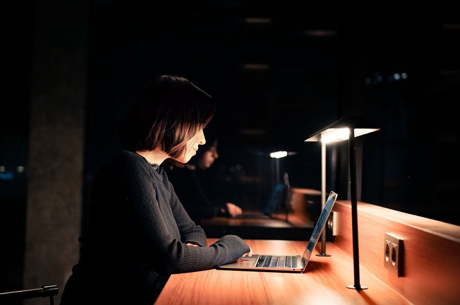 A woman focused on her laptop at a desk with dim lighting, working late at night.