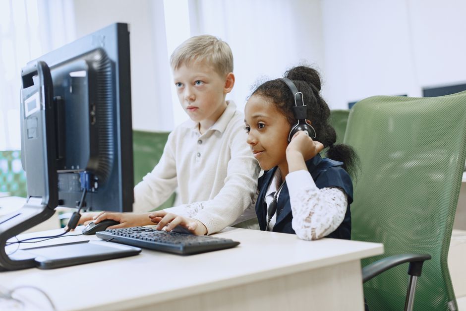 Two children in a computer lab using a desktop computer and headphones.