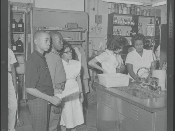 Black students standing in a room with a nurse while other women work next to them in Homer G. Phillips Hospital, on a field trip.