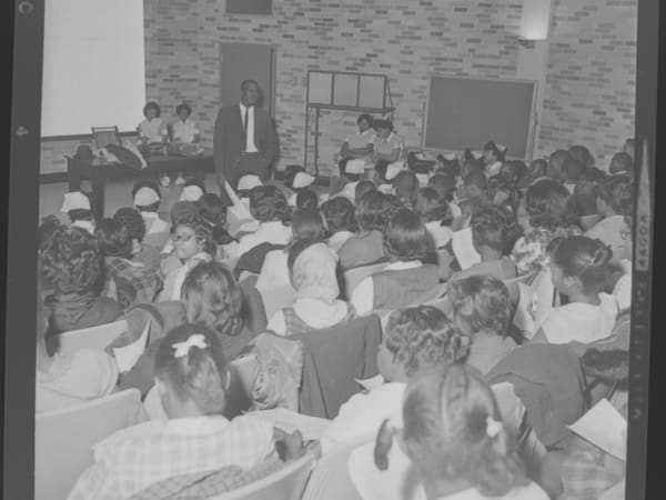 Students sitting in auditorium seating in Homer G. Phillips Hospital, listening to a Black man speak with two Black nurses sitting behind him, as part of a school field trip.