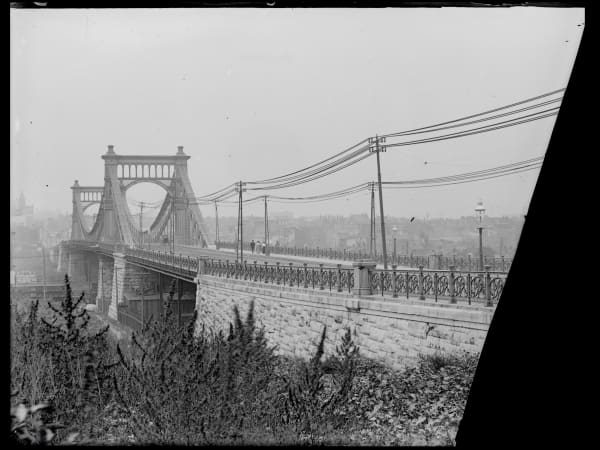 The Grand Avenue suspension bridge from below its stone approach, showing the paired iron towers and braced chains. The glass plate negative is broken at the right.