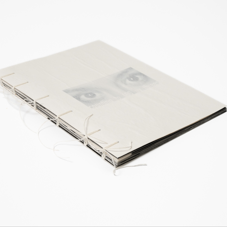 Photo book laid out before white background. A pair of eyes in greyscale are printed on the cover and the title of the piece is below in low-contrast white text. The book is handbound with string.