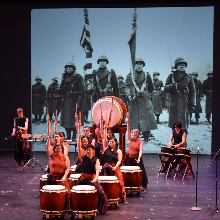 Taiko players pose in front of a black and white photo of Japanese American soldiers in WWII 
