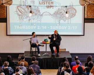 two men speaking into a microphone on stage, recording a podcast