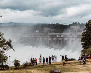 People stand on a river bank observing water flowing over a large dam