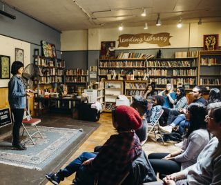 person speaking into a microphone in front of seated audience and shelves of books