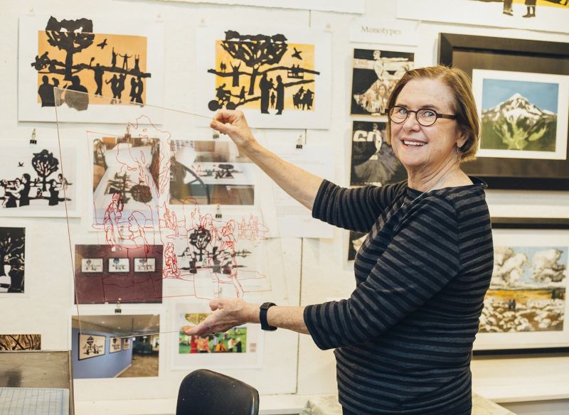 Woman holding up art in front of a wall of hanging paintings