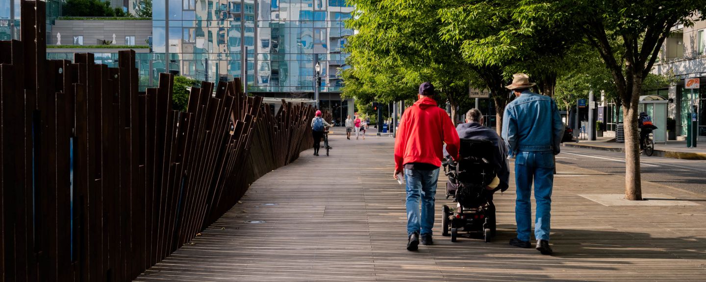 View from behind of a person in red coat and a person in jean jacket walking next to person in a power chair wearing a grey sweatshirt on a wooden walkway. 