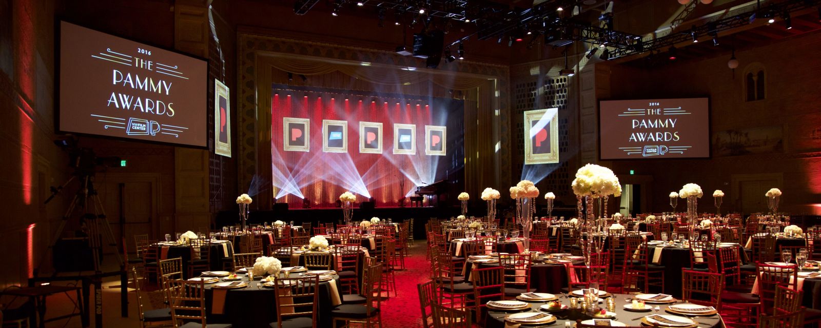 large ballroom with round tables set for dining and a stage in front with screens on each side saying "the Pammy Awards"