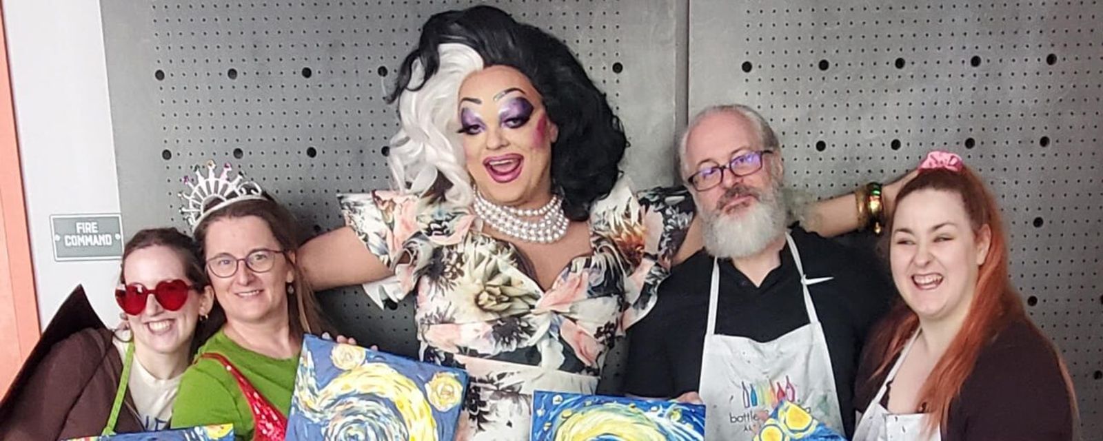 A group of smiling people wearing paint-covered aprons pose with their paintings, standing next to a smiling drag queen