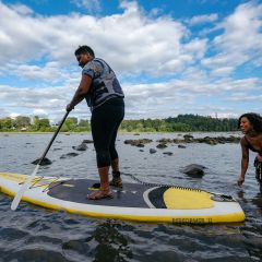 one person wearing a life jacket pilots a stand-up paddleboard in a river while a second person in the water looks on