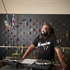 A Black man stands behind a set of two turntables and in front of a black wall displaying dozens of small artworks.