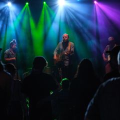 a band plays in front of a crowd under green, blue, and purple stage lights