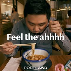 A man pulls noodles out of a bowl with chopsticks in a restaurant under the words \"Feel the ahhh\" and \"Portland\"