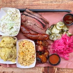 a tray of barbecued meat, sauces, sides of pickle and cucumber, and coleslaw, mashed potatoes and macaroni and cheese