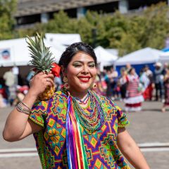 A woman in a colorful embroidered huipil blouse holds a pineapple and smiles