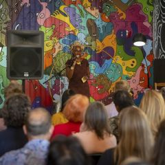 An audience listens to a speaker standing in front of a colorful mural.
