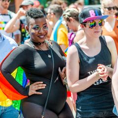 people stand watching a parade wearing rainbow flags and rainbow makeup with rainbows reflecting in their sunglasses