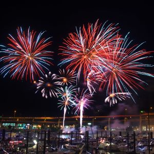 Fireworks burst in a night sky above a river, with boats in the foreground and a bridge in the background