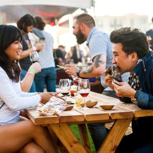 two people eating food and drinking wine at a picnic table during a busy food festival