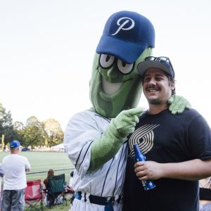 a giant pickle mascot wearing a baseball uniform and hat poses for a photo with a man wearing a Portland Trailblazers T-shirt