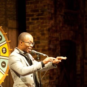 a man speaking on stage with a gameshow wheel behind him