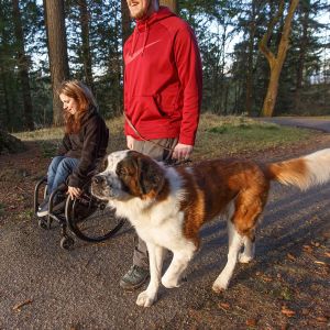 a girl in a wheelchair on nature trail with a man and a dog