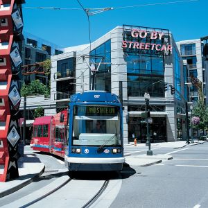 A streetcar passes by a large, red sculpture in front of a building with a neon sign reading, \"Go By Streetcar.\"