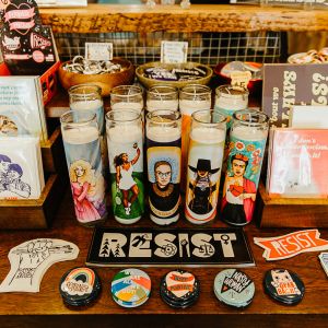 a display of cards, stickers, pins, and votive candles for sale. the items carry slogans like \"support local artists,\" \"feminist,\" \"body positive\" and \"resist.\"