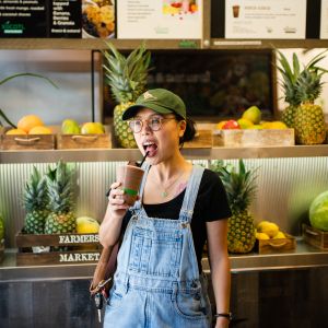 A woman sips a smoothie through a straw from a plastic cup in front of a display of fresh fruit