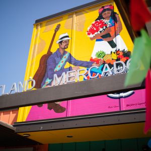 A sign reading \"Portland Mercado\" in front of a brightly painted mural showing a Latin American dancer and musician